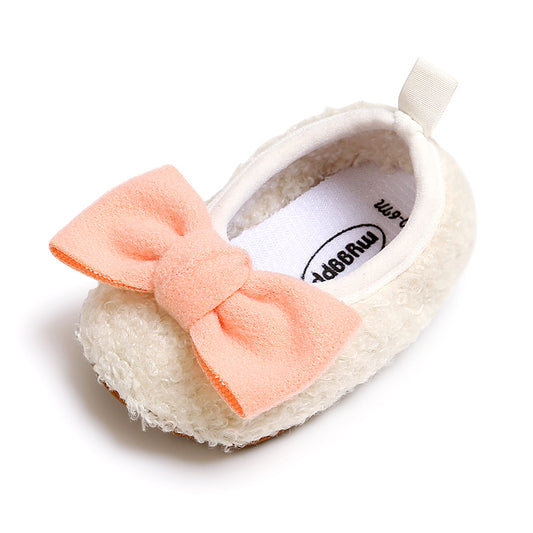 A small, fluffy cream-colored baby shoe with a soft peach bow on top. The shoe has a fabric loop at the back and white interior lining.