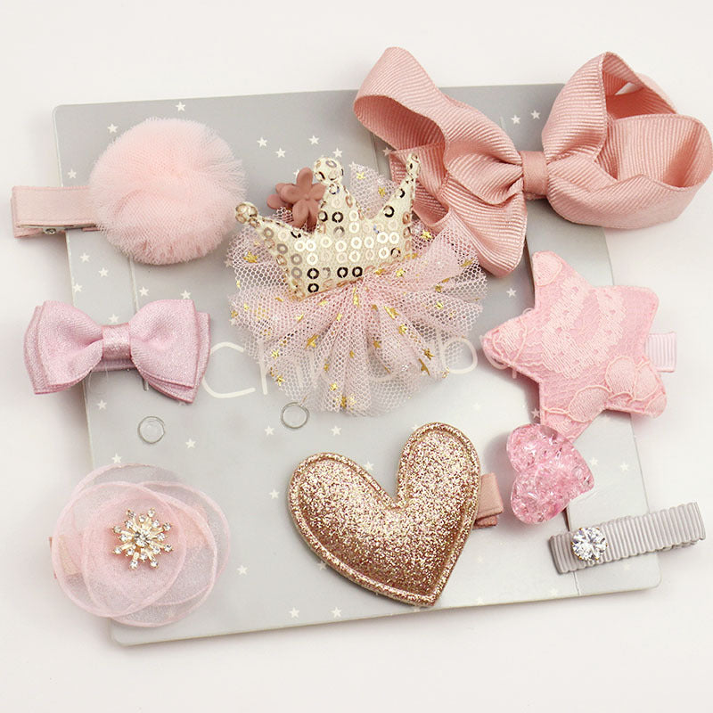 A collection of pink and gold hair accessories on a gray star-patterned background, featuring bows, a sequined crown, a lace star, and a glittery heart.
