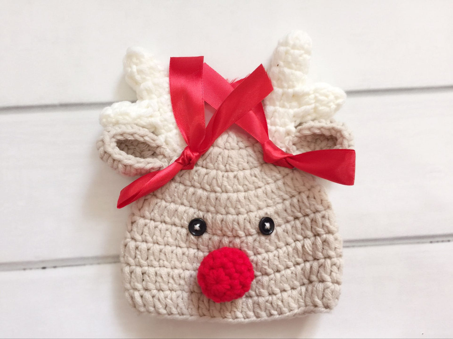 Handmade crochet reindeer hat with antlers, red nose, and button eyes, adorned with a red ribbon, on a white background.