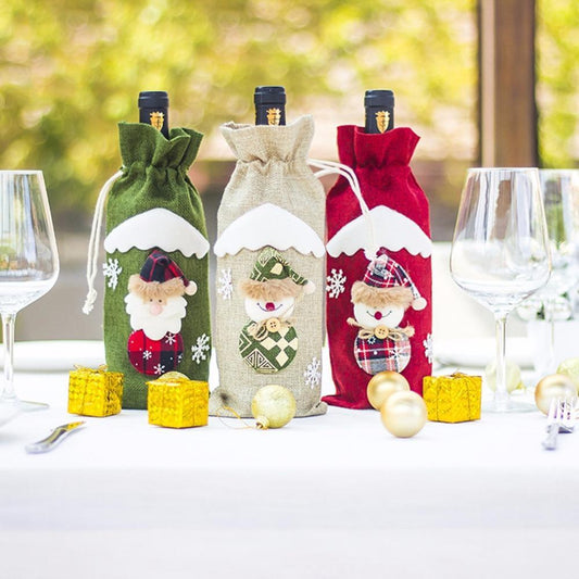 Three wine bottles in festive covers with Santa and snowman designs stand on a table with empty glasses, yellow gift boxes, and gold ornaments against a blurred background.