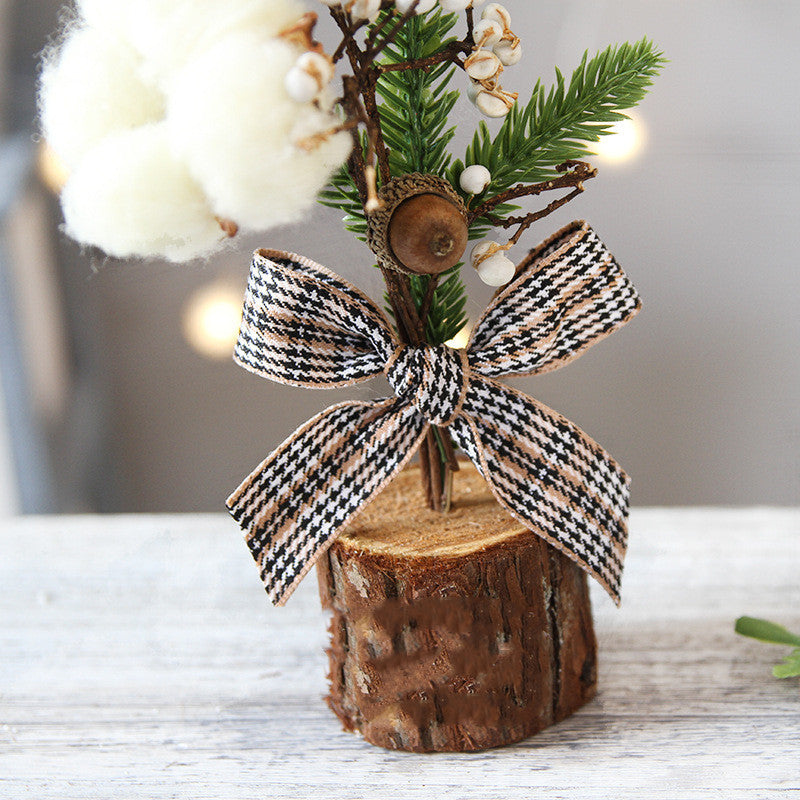 A small decorative arrangement with faux greenery, cotton, and an acorn, set in a wood base, adorned with a plaid bow, on a light wooden surface.