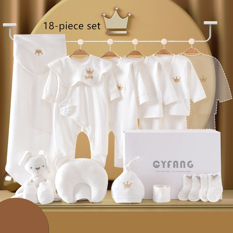 Elegant 18-piece baby gift set features white clothing with crown motifs, plush toys, socks, and accessories. Displayed against a soft gold backdrop with a branded gift box.