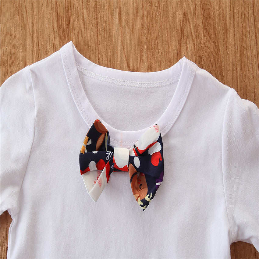 White t-shirt with a colorful bow tie featuring abstract patterns, laid out on a wooden surface. The bow tie includes shades of red, navy, white, and beige.
