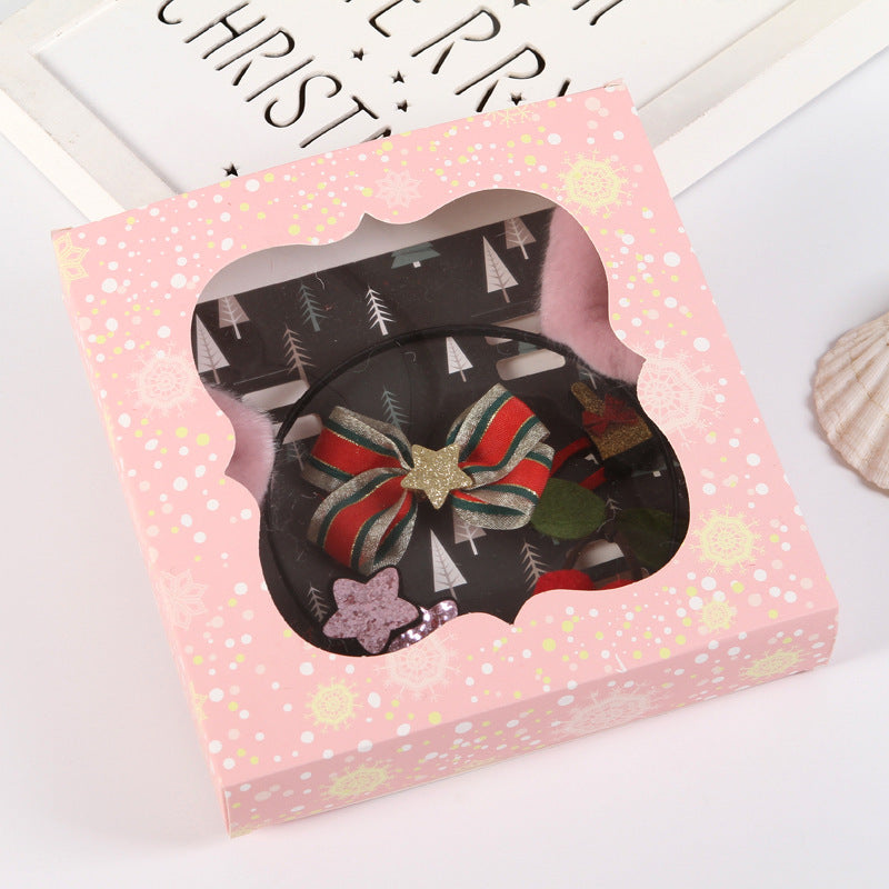 Festive pink gift box with scalloped window displaying a hair accessory featuring a bow with red and green stripes, a gold star, and other decorative elements.
