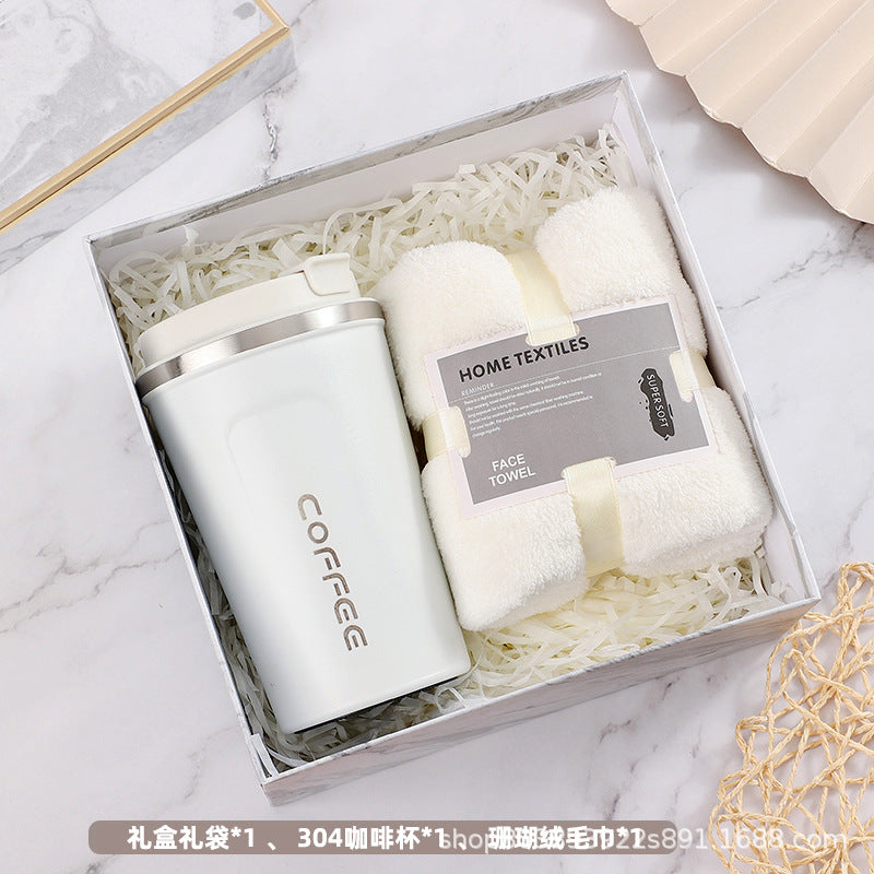 Gift box with a white travel coffee mug labeled "COFFEE" and a soft white face towel labeled "HOME TEXTILES," placed on shredded packing paper.