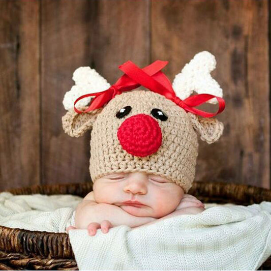 A sleeping baby wears a handmade reindeer hat with a red nose, white antlers, and a red bow, nestled in a basket against a wooden background.