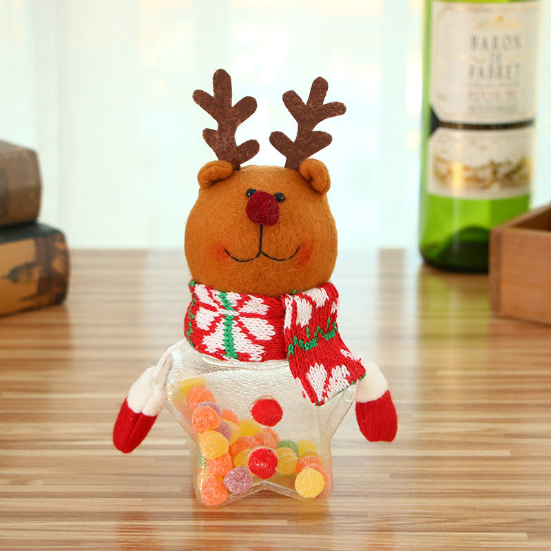 A cute reindeer figure with antlers and a red scarf sits on a wooden table. Its body is a star-shaped jar filled with colorful candy. A green wine bottle is in the background.