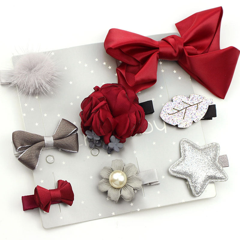 A set of seven hair accessories featuring bows, pom-poms, and floral designs in shades of red, gray, and glittery silver, displayed on a star-patterned white background.