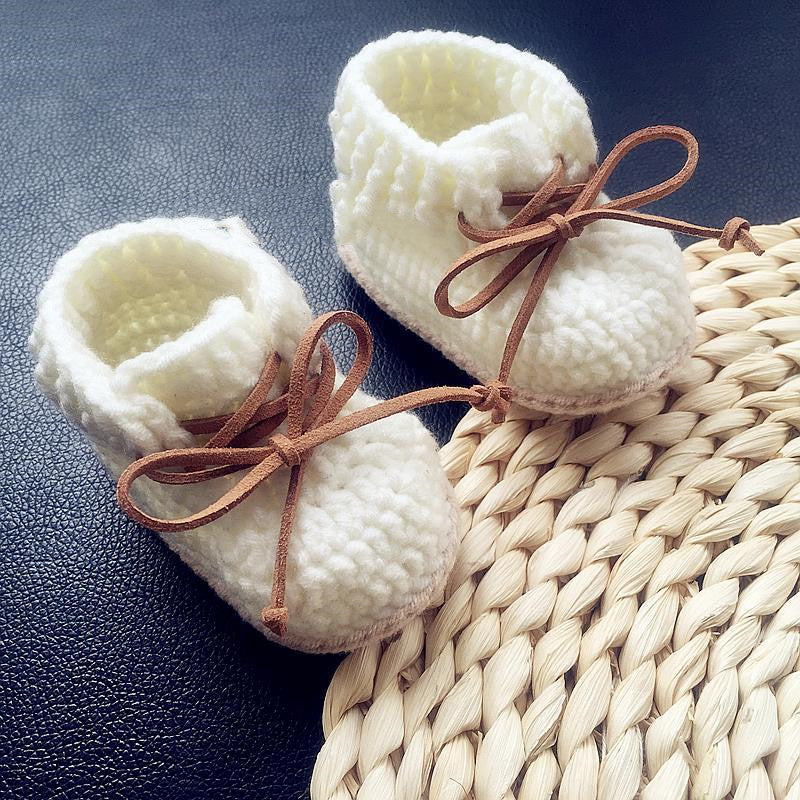 White knitted baby booties with brown laces, placed on a black leather surface, partially on a woven beige coaster.
