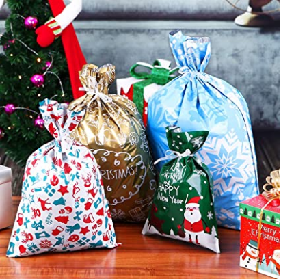 Four festive gift bags, decorated with Christmas and New Year designs, are placed on a wooden table. A Christmas tree and small gift boxes are in the background.