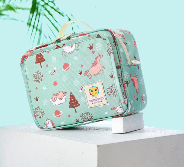 Green children's bag with cartoon animals and trees, featuring a top handle. It rests on a white block with a palm frond in the background, set against a pastel blue backdrop.