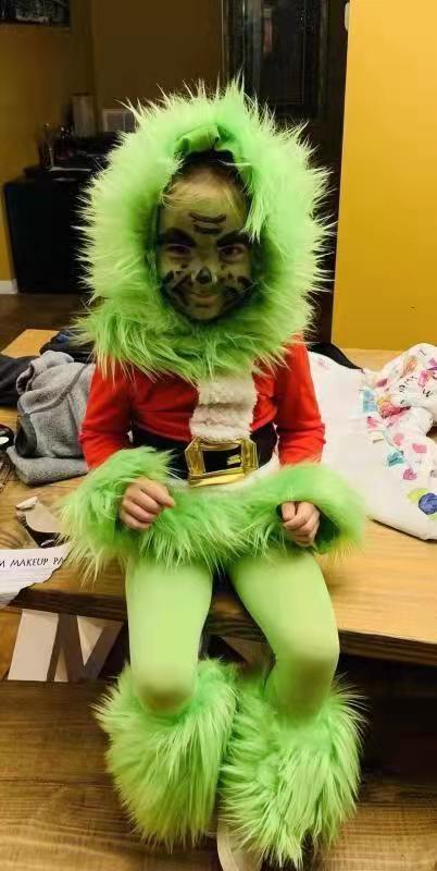 Child dressed as a festive green creature with fluffy fur, face paint, red top, and green leggings, sitting on a bench in a cozy room with a wooden floor.