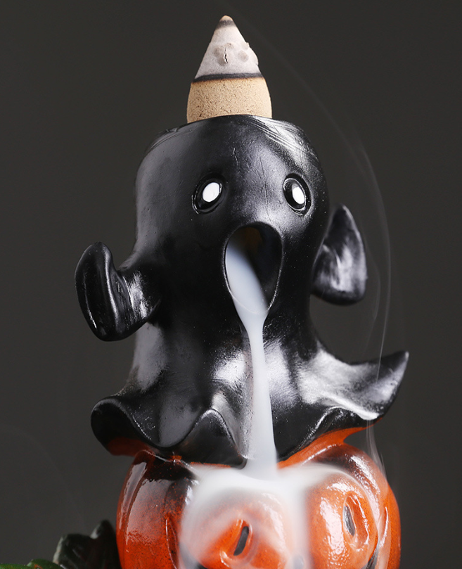 A black ceramic ghost incense holder featuring a cone on top, releasing white smoke that flows through its open mouth and down onto a carved orange pumpkin base.