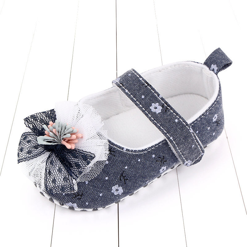 Navy blue baby shoe with floral patterns, featuring a strap and a decorative bow with lace and pastel flower accents, set on a white wooden floor.