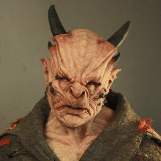 A creature with a wrinkled, pinkish face, dark horns, and black markings, wearing a tattered garment. The expression is menacing against a plain background.