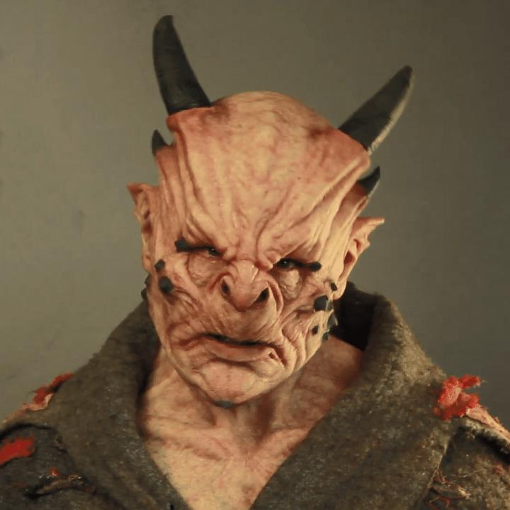 Detailed demon-like creature with textured skin, horns, and a fierce expression, wearing a tattered cloak with red accents. The background is plain.