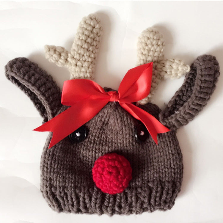 Knitted reindeer hat with antlers and red pompom nose, featuring black button eyes and a decorative red bow on top, set against a white background.