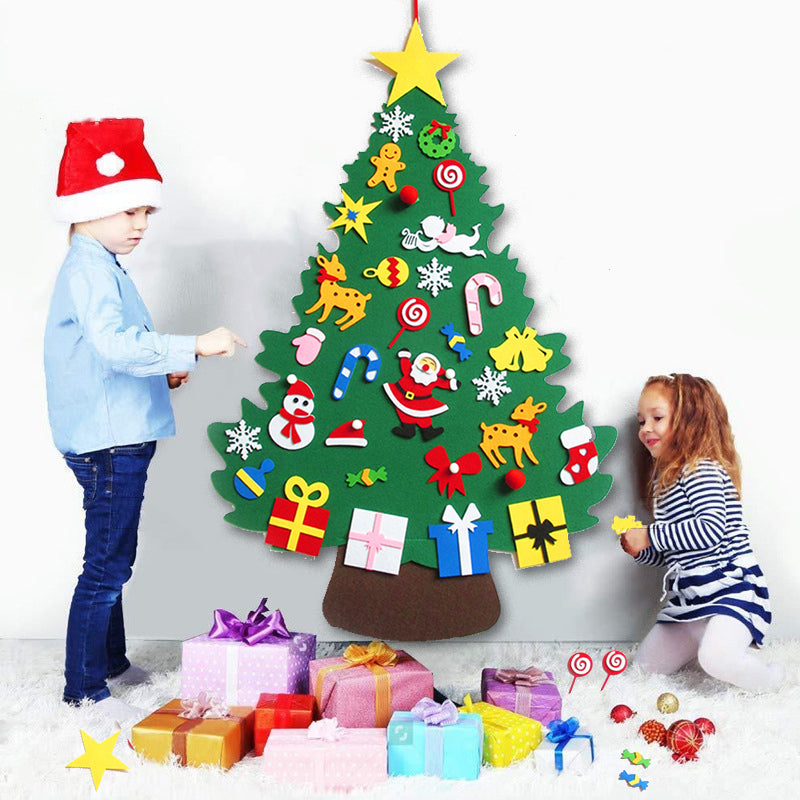 Two children decorate a felt Christmas tree with colorful ornaments. A boy in a Santa hat stands on the left, while a girl in stripes sits on the right. Gifts are piled underneath.