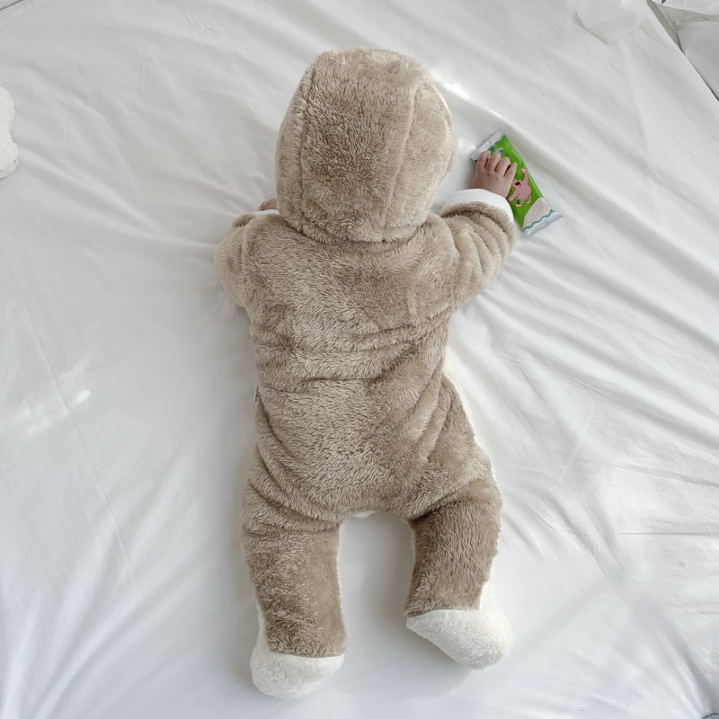 A baby in a plush, brown bear onesie with white feet lies on a white bed, reaching for a colorful book. The scene is cozy and playful.