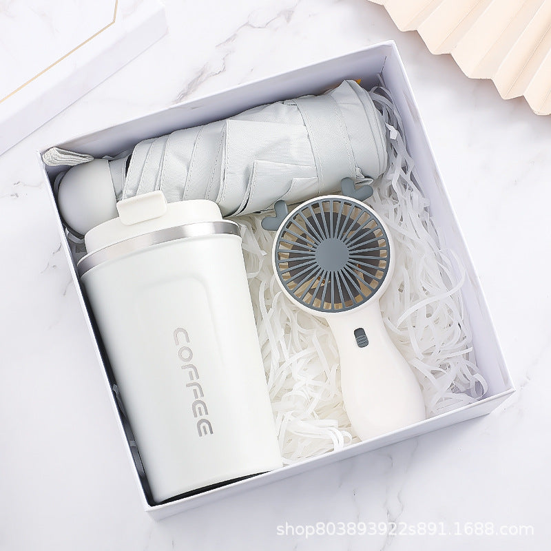 A gift box containing a folded light gray umbrella, a white coffee tumbler labeled "COFFEE," and a small handheld fan, all nestled in shredded white paper.