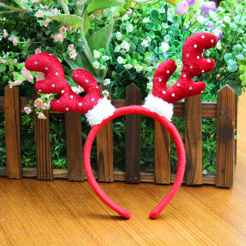 Red reindeer antler headband with white polka dots and fluffy bases, resting on a wooden table. Background of wooden fence and lush greenery with small pink flowers.