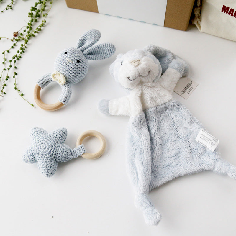 Soft blue and white baby toys on a table, including a crocheted rabbit and star with wooden rings, and a plush bunny comfort blanket. Green plant strands decorate the background.