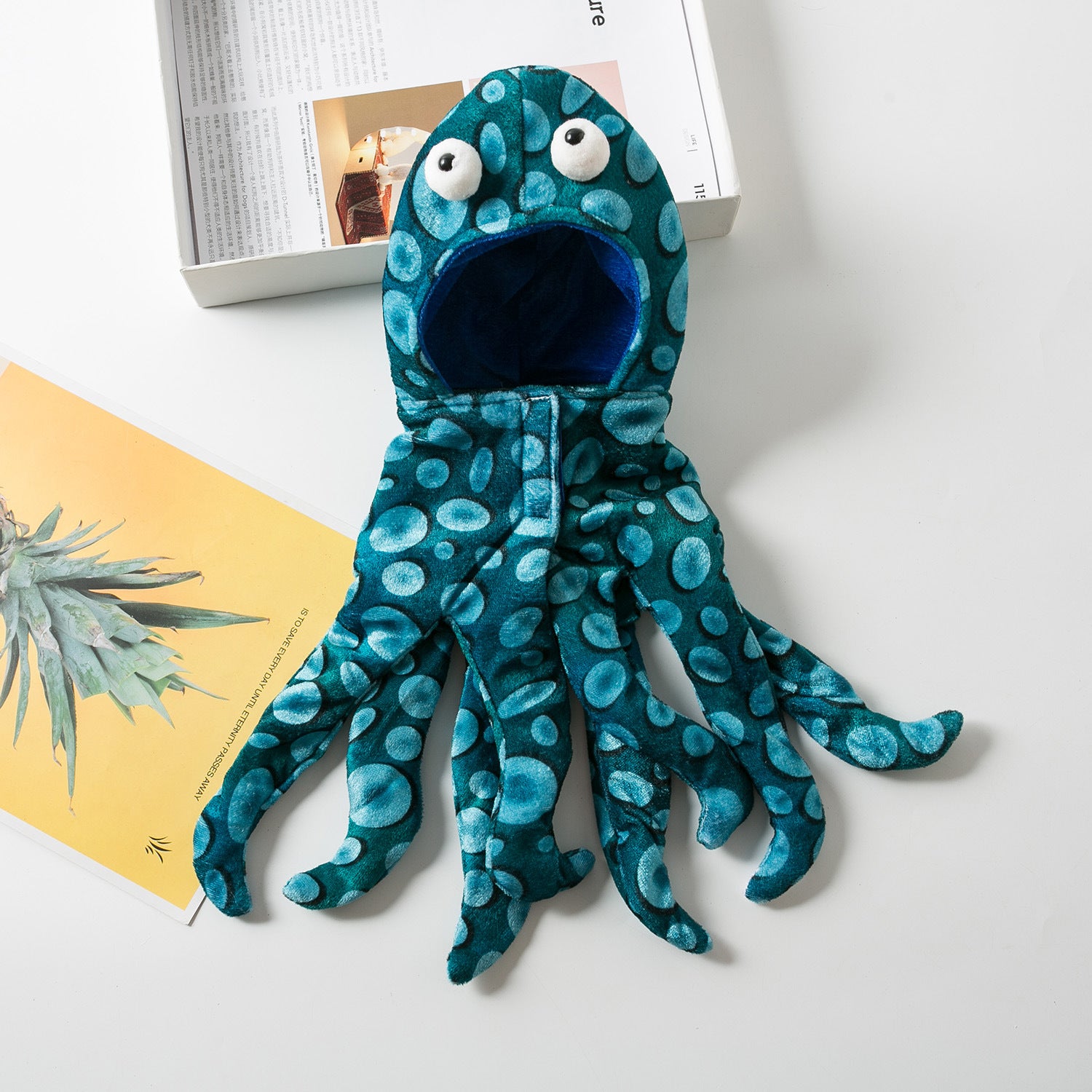 Blue polka-dotted octopus-shaped plush toy with large googly eyes and an open mouth. It's laid out on a white surface near a book and a yellow card with a plant image.