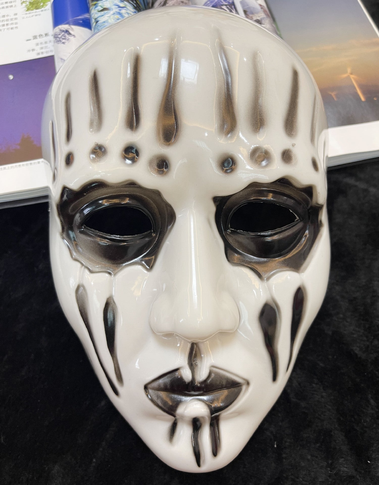 A white theatrical mask with black streaks and patterns around the eyes, forehead, and mouth, lying on black fabric with open book pages visible in the background.