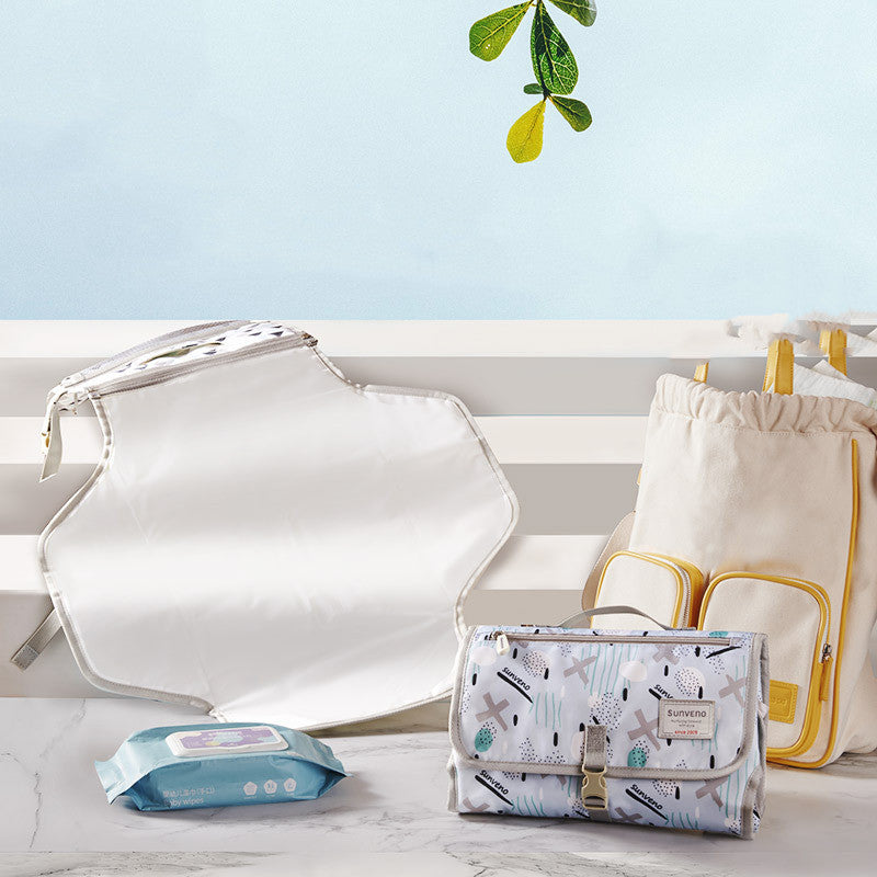 Baby changing kit includes a patterned changing mat, white and yellow diaper bag, and a pack of wipes, all set against a blue background with hanging green leaves.
