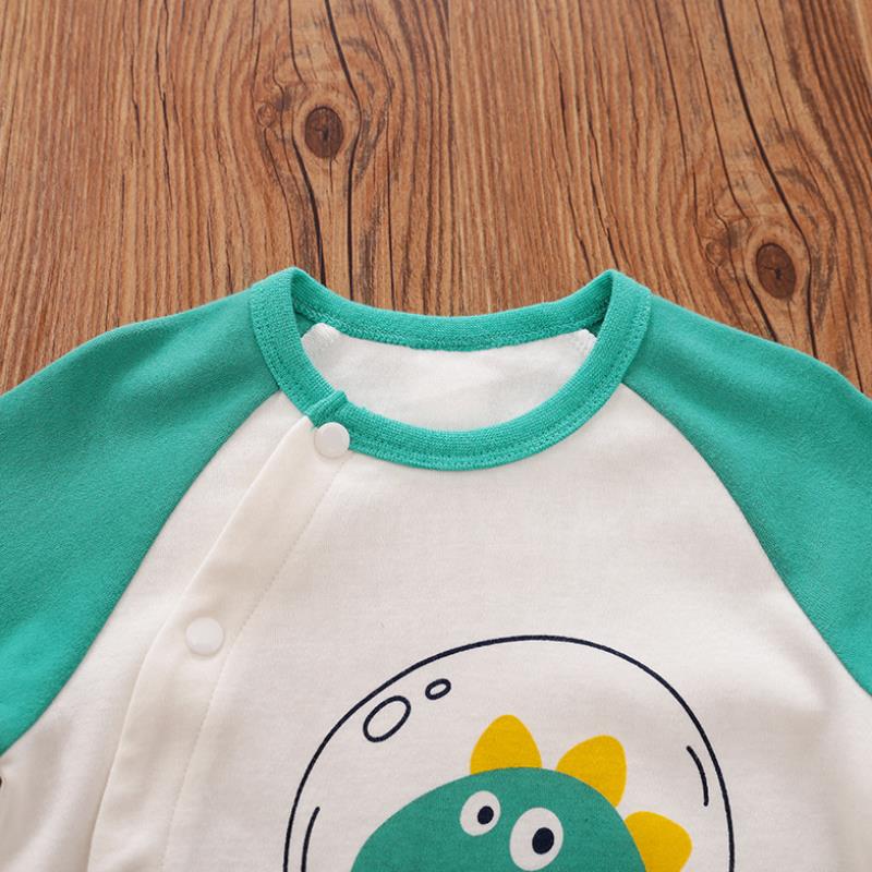 Close-up of a baby onesie with green sleeves, featuring a cartoon dinosaur in a bubble on a white fabric. Snap buttons run along the front. Placed on a wooden surface.