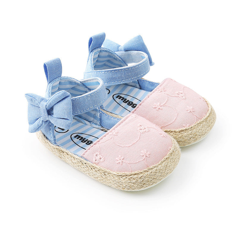 Pink baby sandals with floral embroidery, blue ankle straps, and bows. The soles are woven with a straw-like texture. The interior lining is blue and white striped.