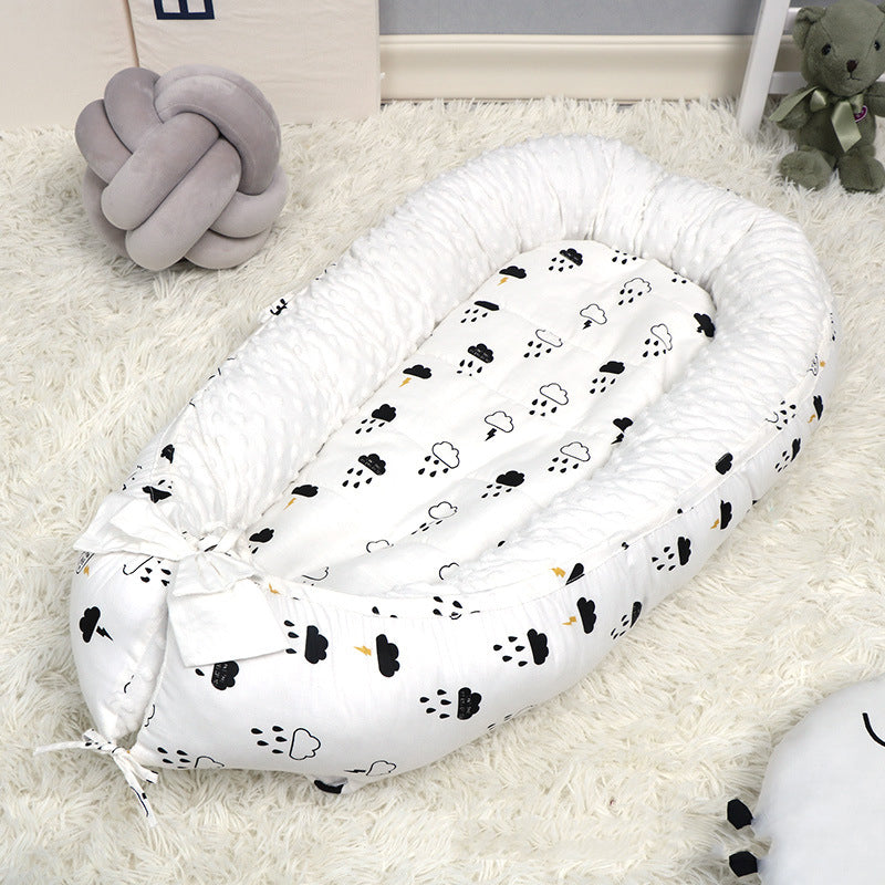 A cozy baby nest with a white and black cloud pattern, placed on a fluffy white rug. Nearby are a gray plush knot pillow and a green stuffed bear on the floor.