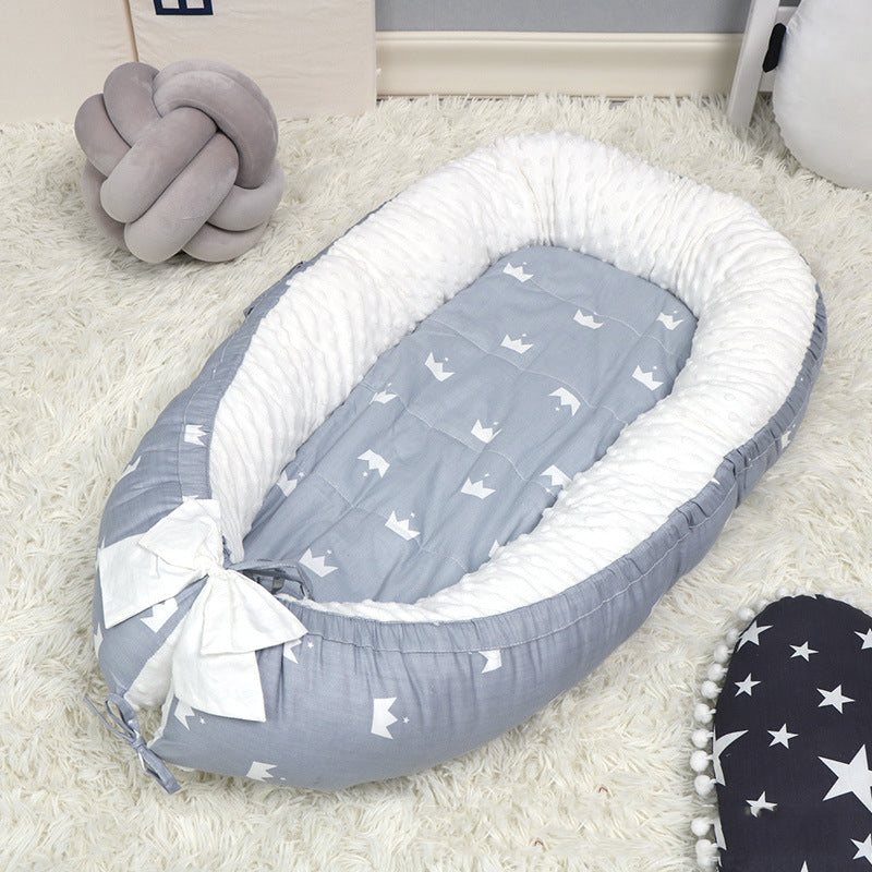 Cozy baby nest with gray and white crown pattern on a soft rug, featuring a white ruffled lining and a bow detail. Nearby are plush decor and a star-patterned cushion.