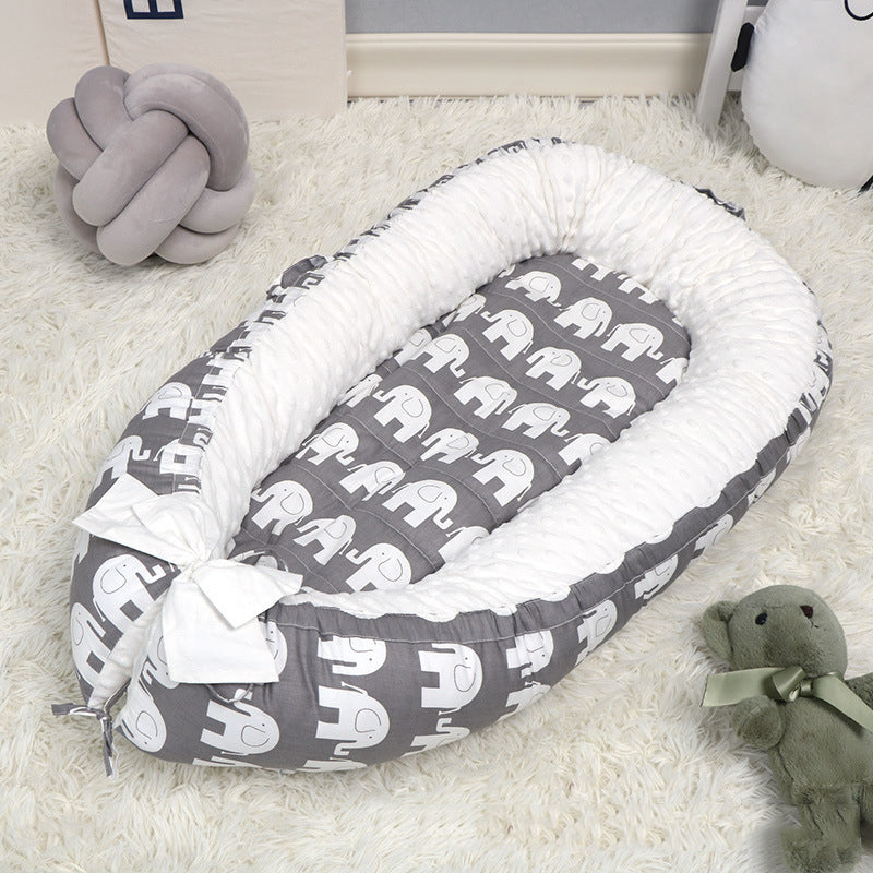 Gray baby nest with white elephant patterns and cushioned edges on a plush rug. A gray knot pillow and green teddy bear are nearby for a cozy nursery setting.