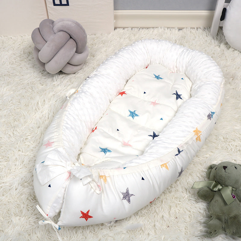 A cozy white baby nest with colorful star patterns is placed on a fluffy rug. Nearby are a soft, knotted cushion and a green teddy bear with a ribbon.