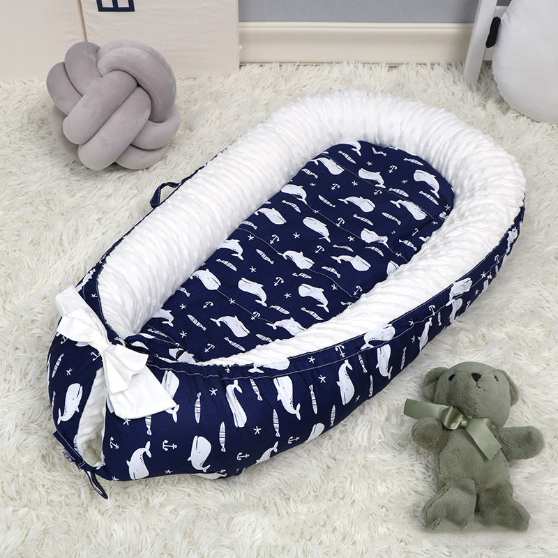 A cozy navy baby nest with white whale patterns on a fluffy rug. It features a white inner lining and a bow, with a gray knot pillow and a green teddy bear nearby.