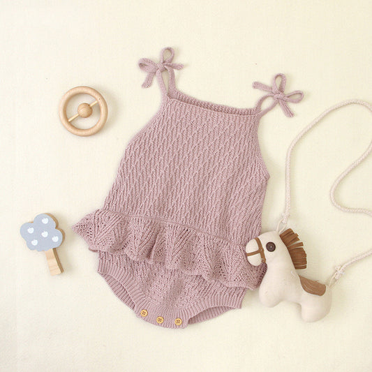 Knitted pink baby romper with shoulder ties, laid on a light surface. Accompanied by a wooden rattle, blue cloud toy, and a small plush horse on a string.