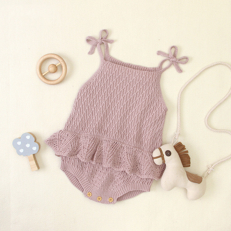 Pink knitted baby romper with a ruffled waist and shoulder ties, surrounded by a wooden rattle, a blue cloud-shaped toy, and a small toy horse with a string.