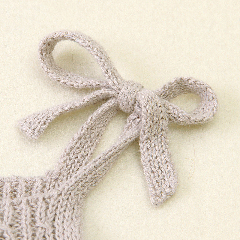 A close-up of a light beige knitted bow made from soft yarn, displayed on a matching fabric surface. The bow is neatly tied with a textured pattern.