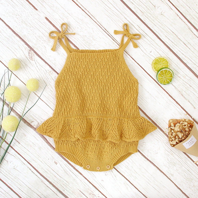 Yellow knitted baby romper with frilled waist and shoulder bows on a wooden surface. Accompanied by decorative yellow pom-poms, sliced citrus, and a popcorn cone.