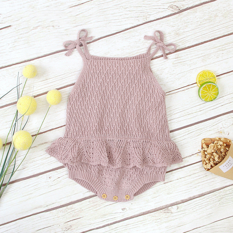 Pink knitted baby romper with ruffled hem and bow straps, displayed on wood surface. Surrounding items include yellow decorative spheres, sliced citrus, and a small basket.