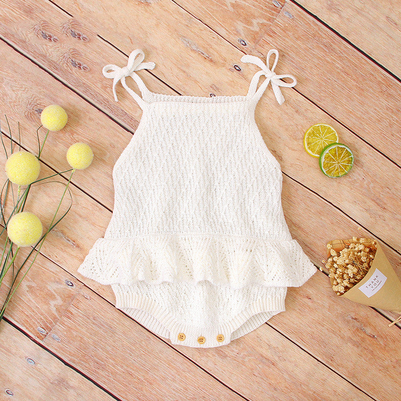 White knitted baby romper with tie straps and ruffled waist on wooden surface. Accompanied by yellow spherical decorations, dried flowers, and sliced limes.