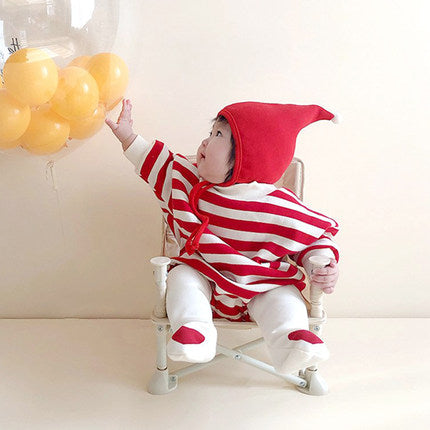 A baby in a red and white striped outfit with a matching hat sits in a small chair, reaching for a bunch of yellow balloons against a light-colored background.