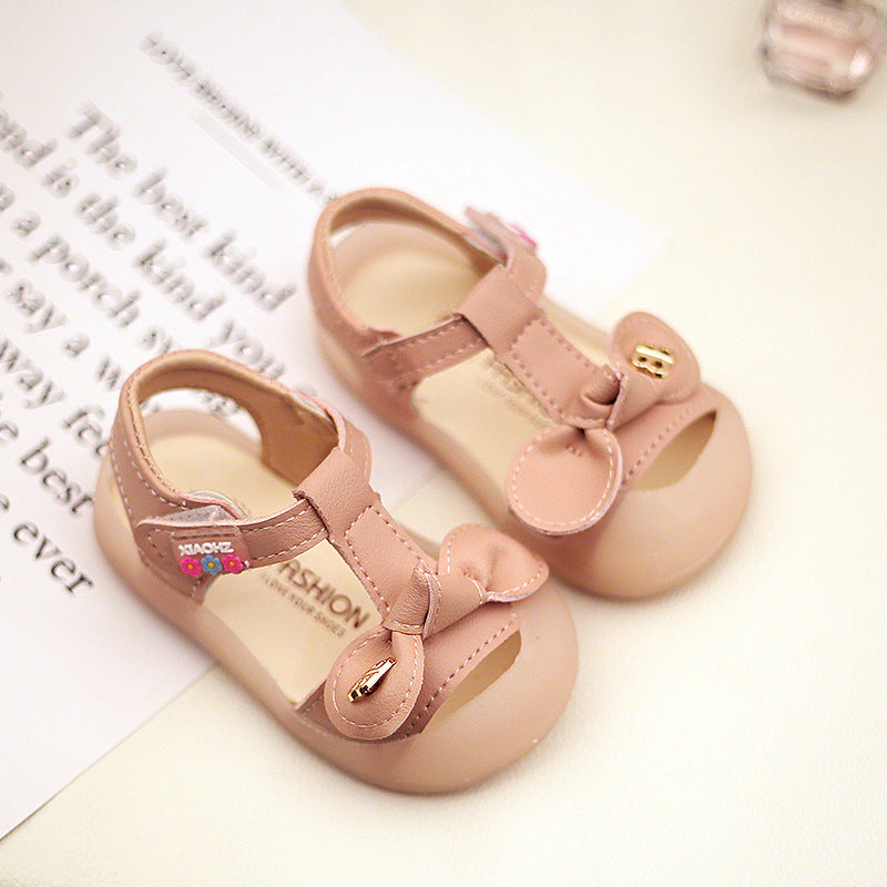 Pink baby sandals with bows and gold accents, placed on a white surface over a piece of paper with text. The sandals have a T-strap design and decorative floral patches.