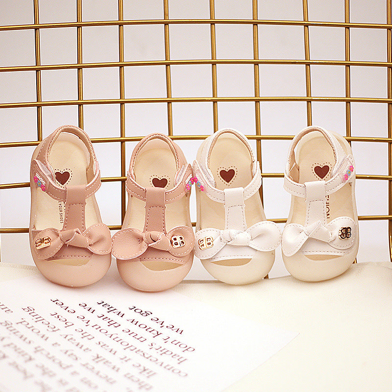 Four baby shoes displayed on a grid backdrop. Two are pink and two are white, each featuring a bow and heart details on the insole with gold accents.