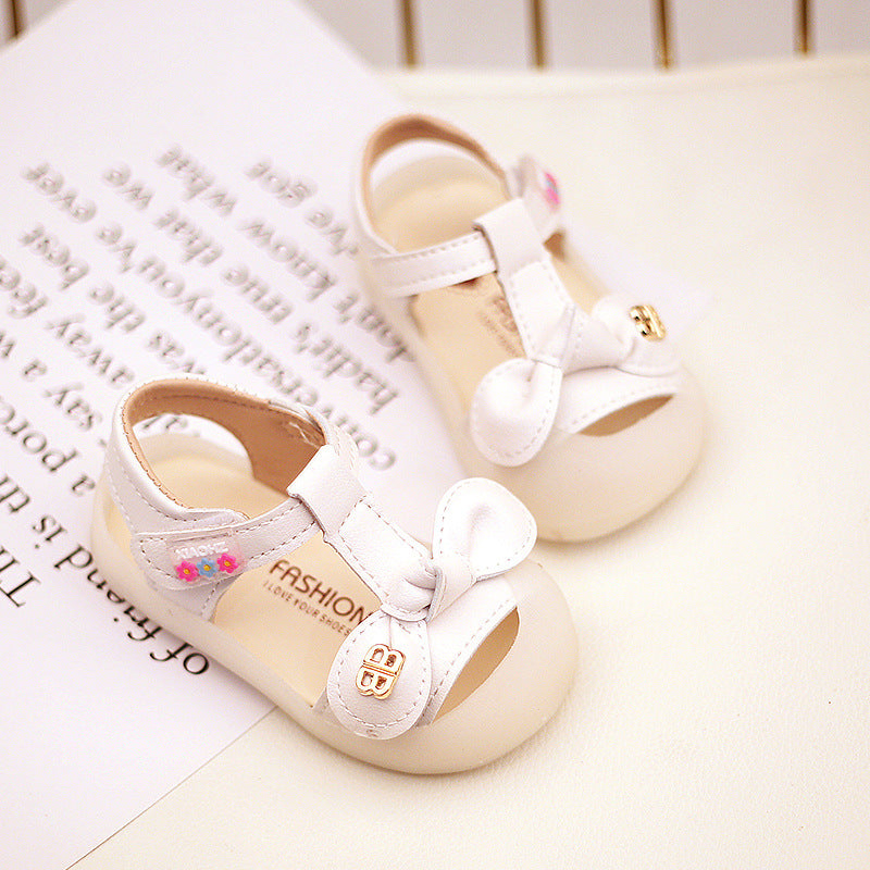 White baby sandals with a bow detail and a small button accent. The shoes have open toes and are placed on a paper with blurred text in the background.