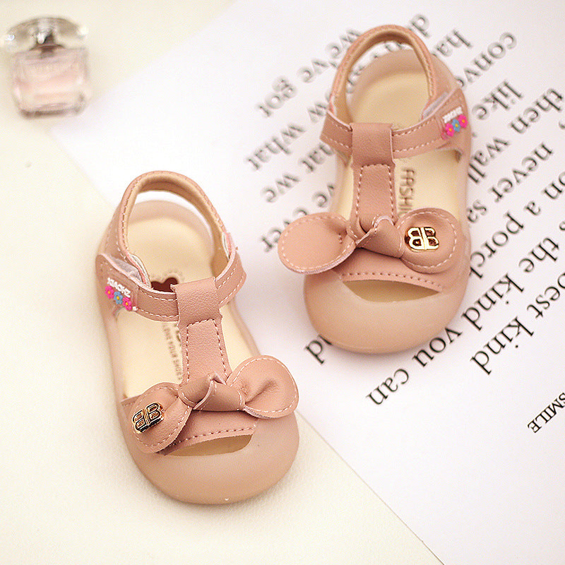 Beige baby shoes with ankle straps and bow accents on a white surface with writing. A small pink bottle is in the background.