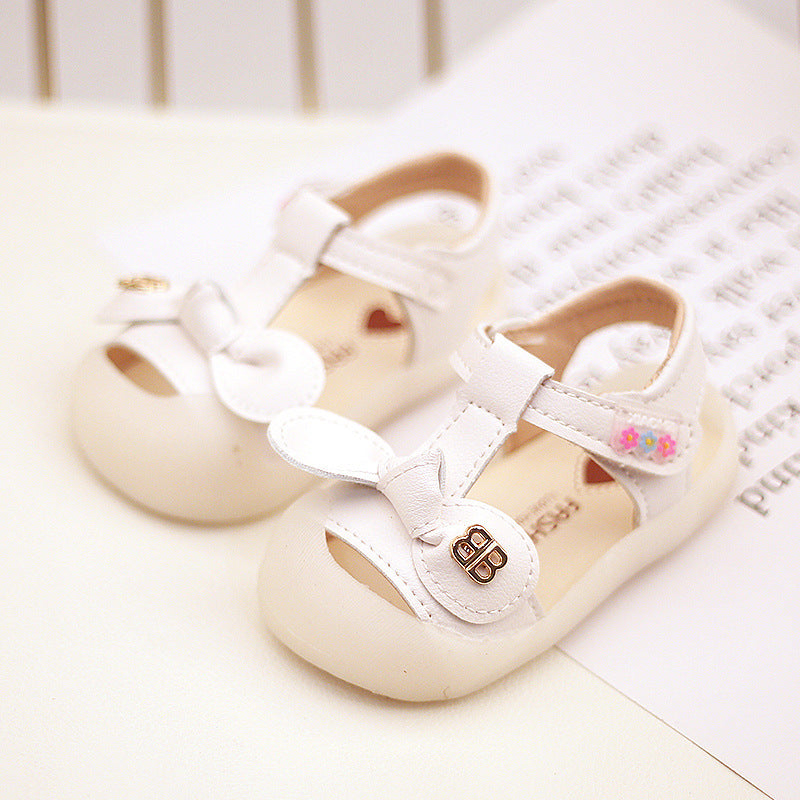 Two white toddler sandals with soft soles and closed toes. They feature a bow detail, floral accents on the straps, and metallic embellishments, placed on a white surface.