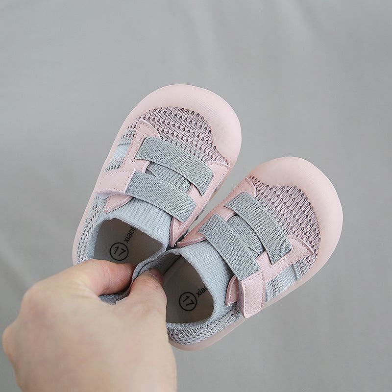 A hand holds a pair of small, pink toddler shoes with grey mesh and adjustable straps, showing size label “17” on the insoles. The background is a neutral grey.