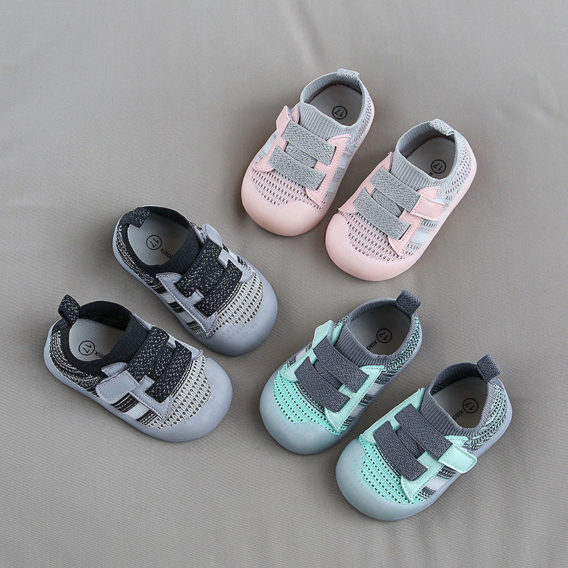 Three pairs of baby shoes are arranged on a gray surface: pink and gray, black and gray, and turquoise and gray. They have soft fabric tops and elastic bands.